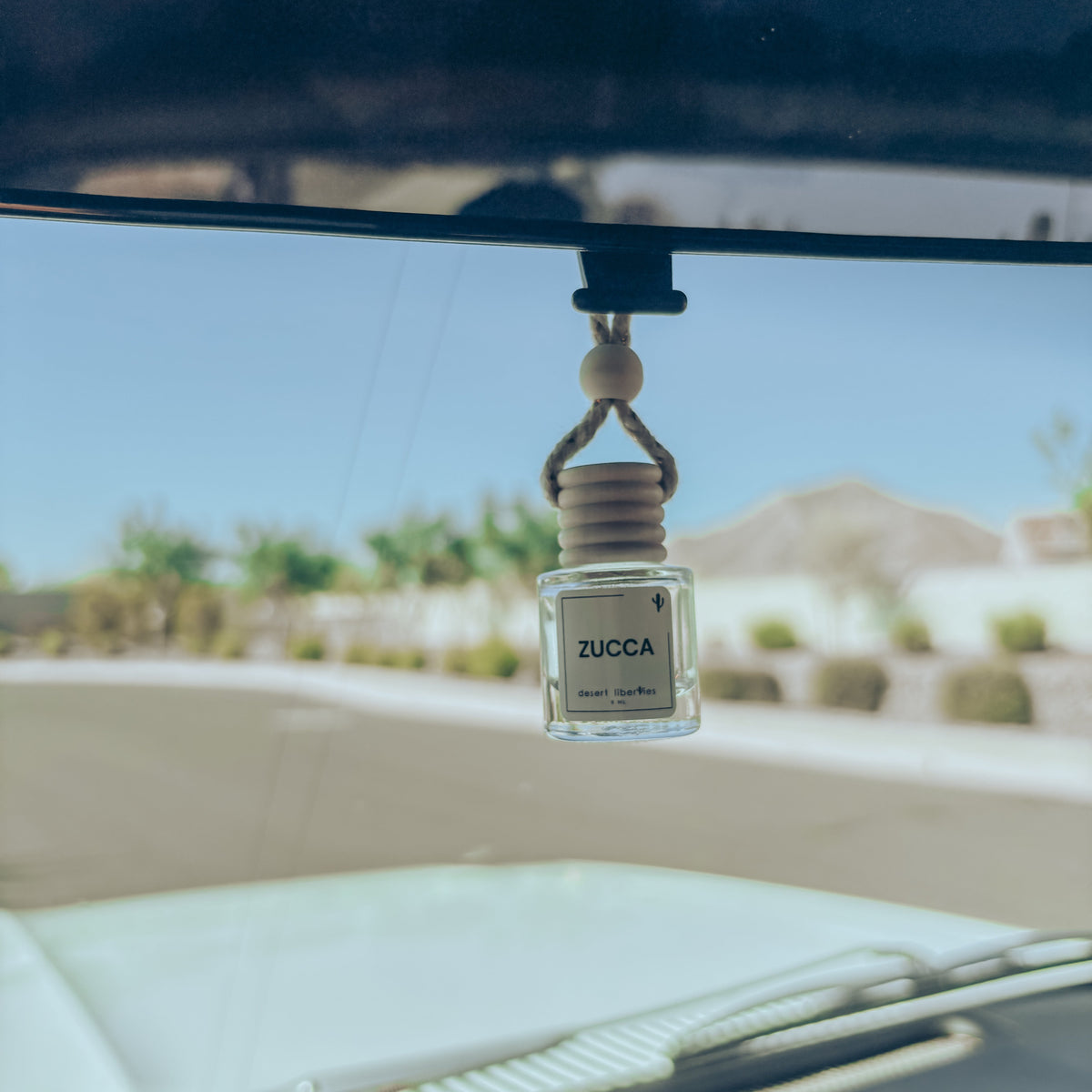 Zucca car air freshener hanging from a car's rearview mirror with a desert landscape in the background.