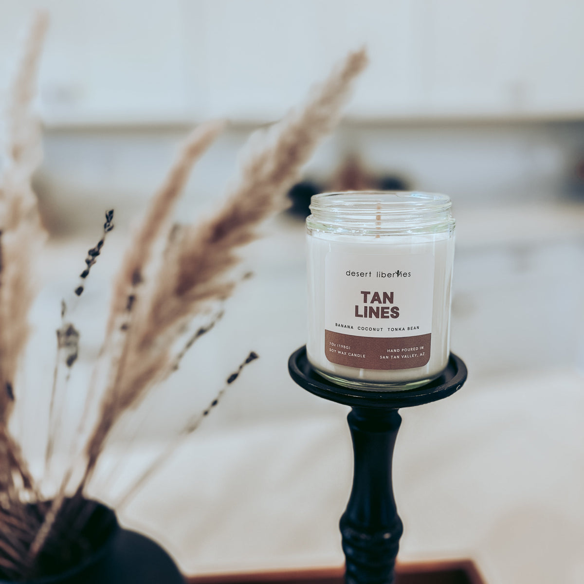 Candle labeled 'Tan Lines' on a black holder with dried plants in the background