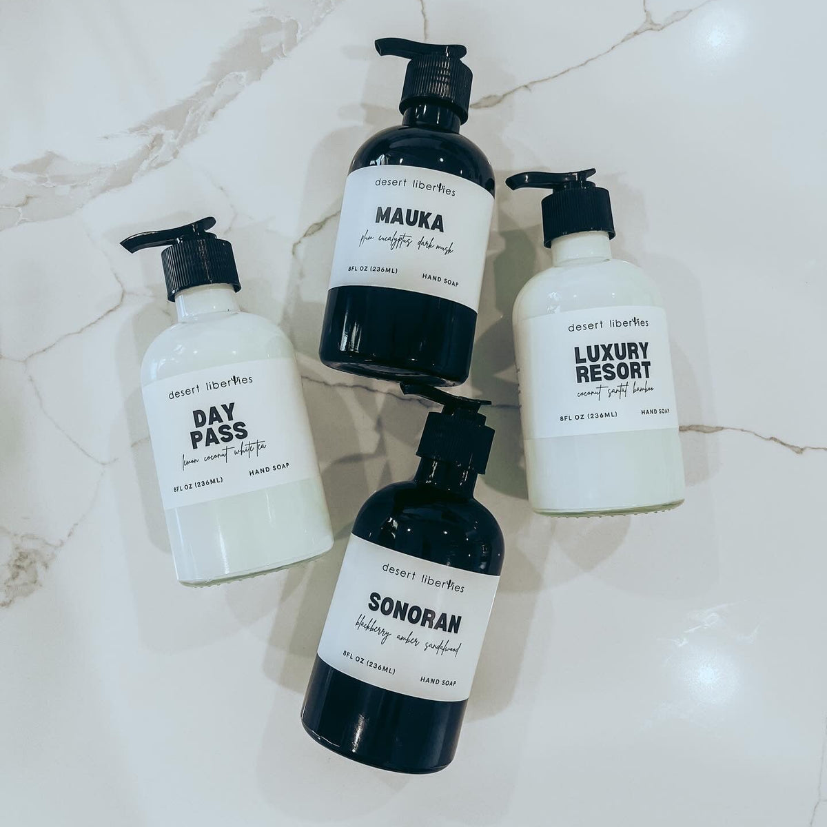 Four reusable glass bottles of organic hand soap with black pump dispensers, labeled with different scents such as 'DAY PASS', 'MAUKA', 'LUXURY RESORT', and 'SONORAN'. Two bottles are white and two are black, displayed on a marble surface.