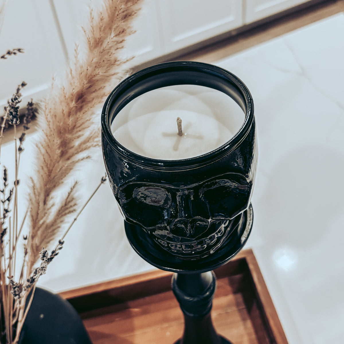 A black skull-shaped candle holder candle with white wax, displayed on a wooden tray next to dried pampas grass in a home setting.