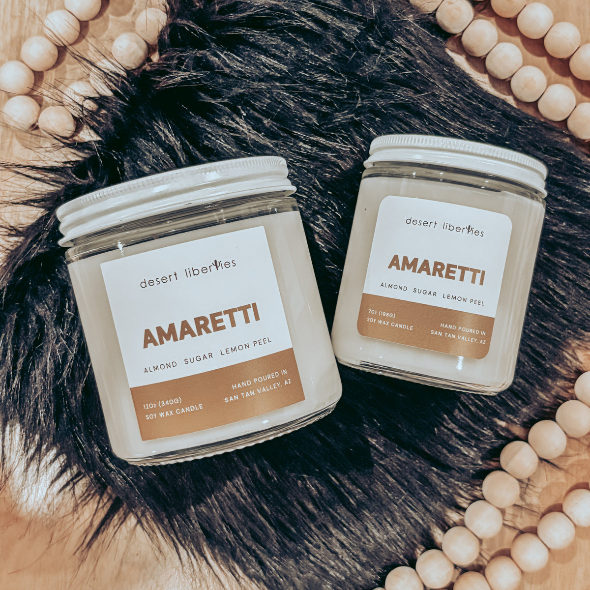 Two 'Amaretti' candles from Desert Liberties on a wooden surface with decorative beads.