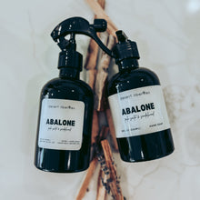 Load image into Gallery viewer, Two black bottles labeled 'Abalone' on a marble surface with palo santo sticks.