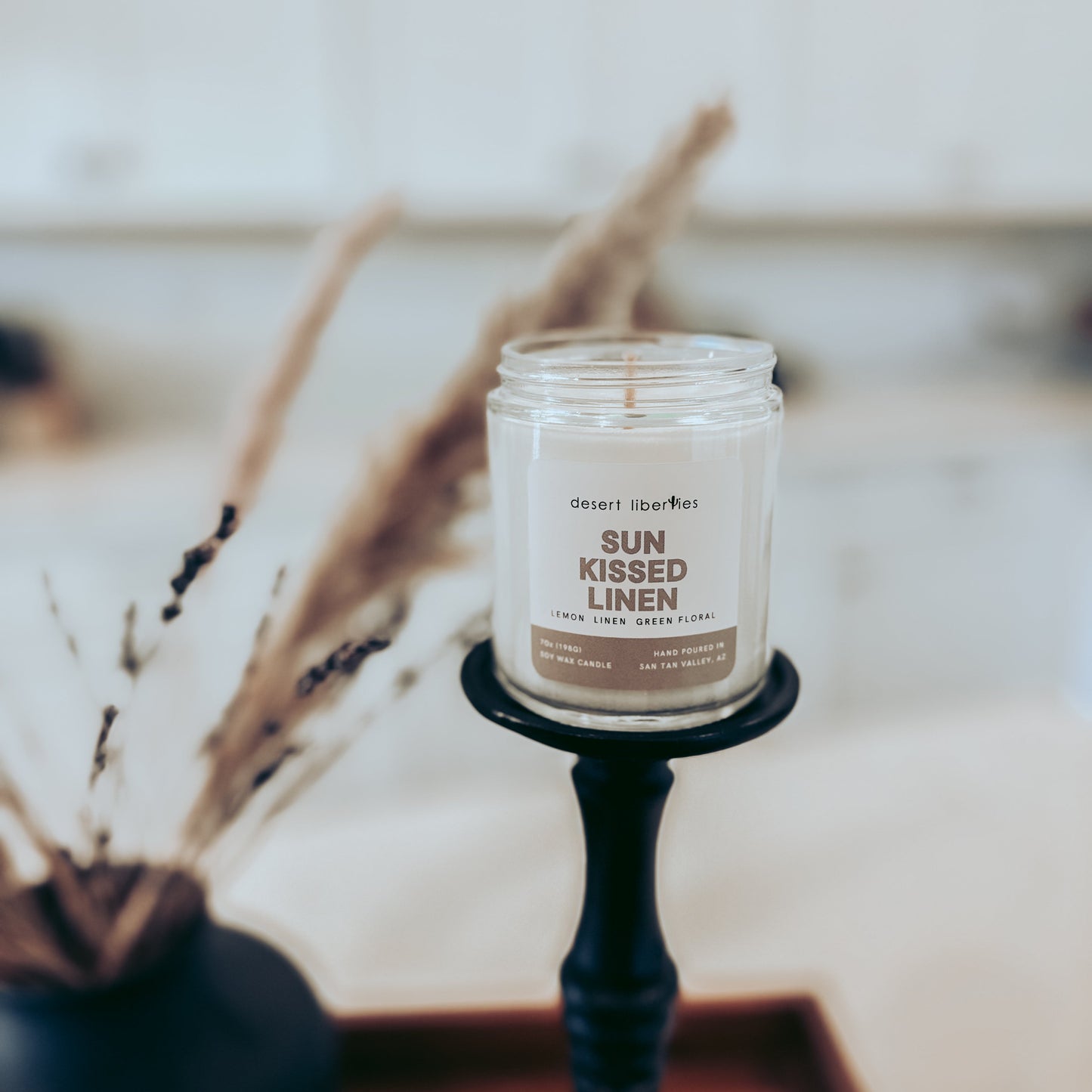 Candle labeled 'Sun Kissed Linen' on a black holder with dried plants in the background