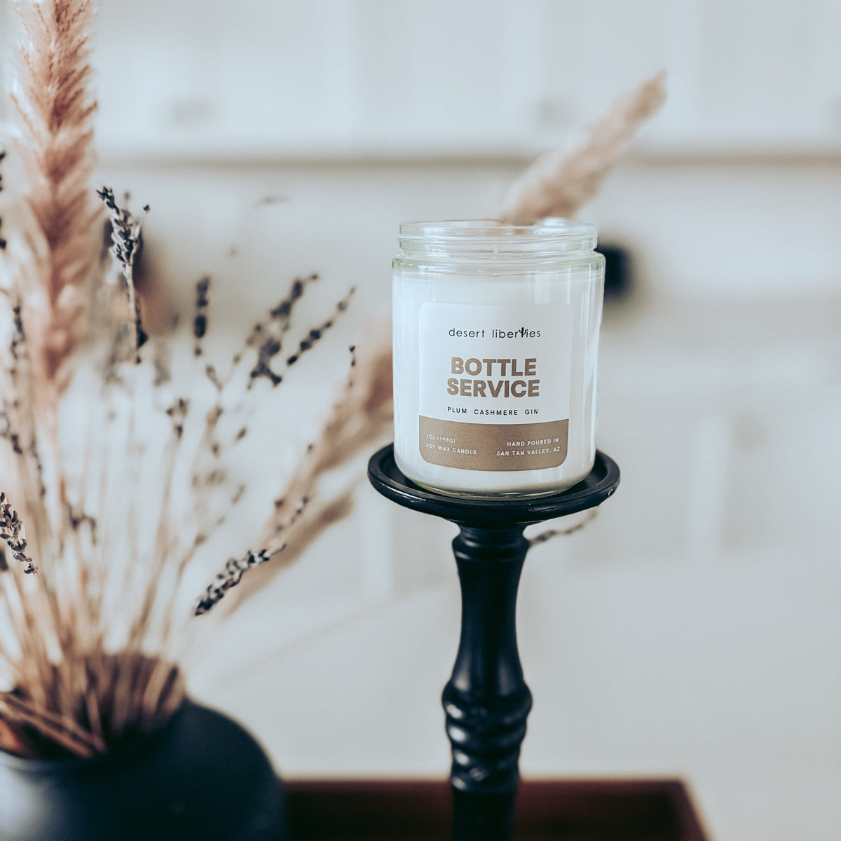 A white jar candle labeled 'BOTTLE SERVICE' sits on a black candle holder, styled with dried grasses in a dark vase against a softly blurred white background.