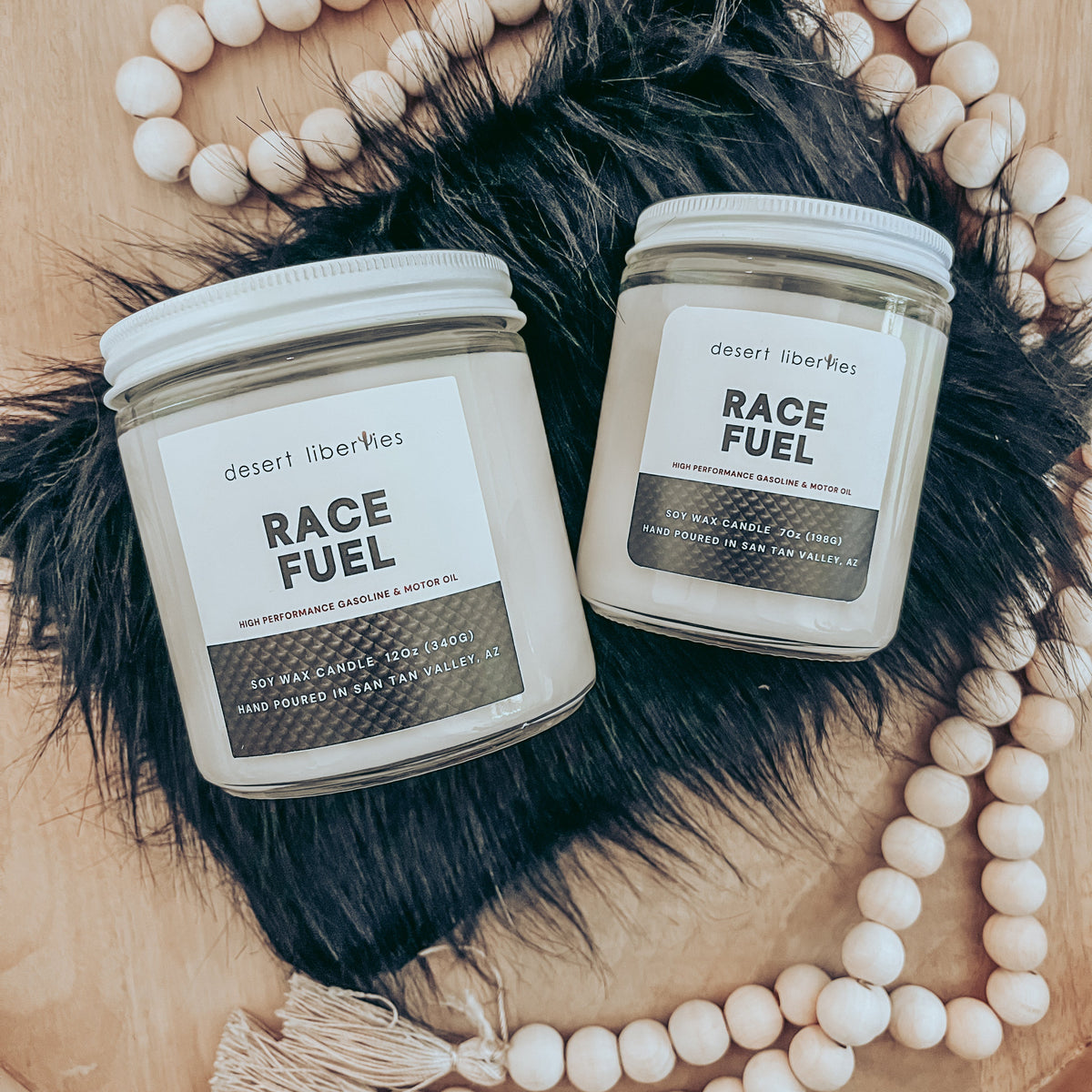 Two candles labeled 'RACE FUEL' by Desert Liberties, with carbon fiber-like label, placed on a wooden surface with a black fur accent and beads, with a shirt in the background.
