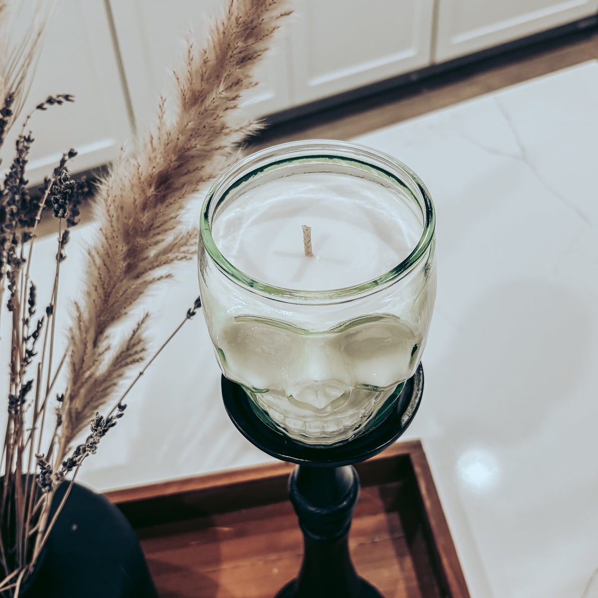 A clear glass jar candle with a wick sits on a black pedestal holder on a wooden tray, with dried pampas grass and a dark vase in the foreground and white cabinetry in the background.