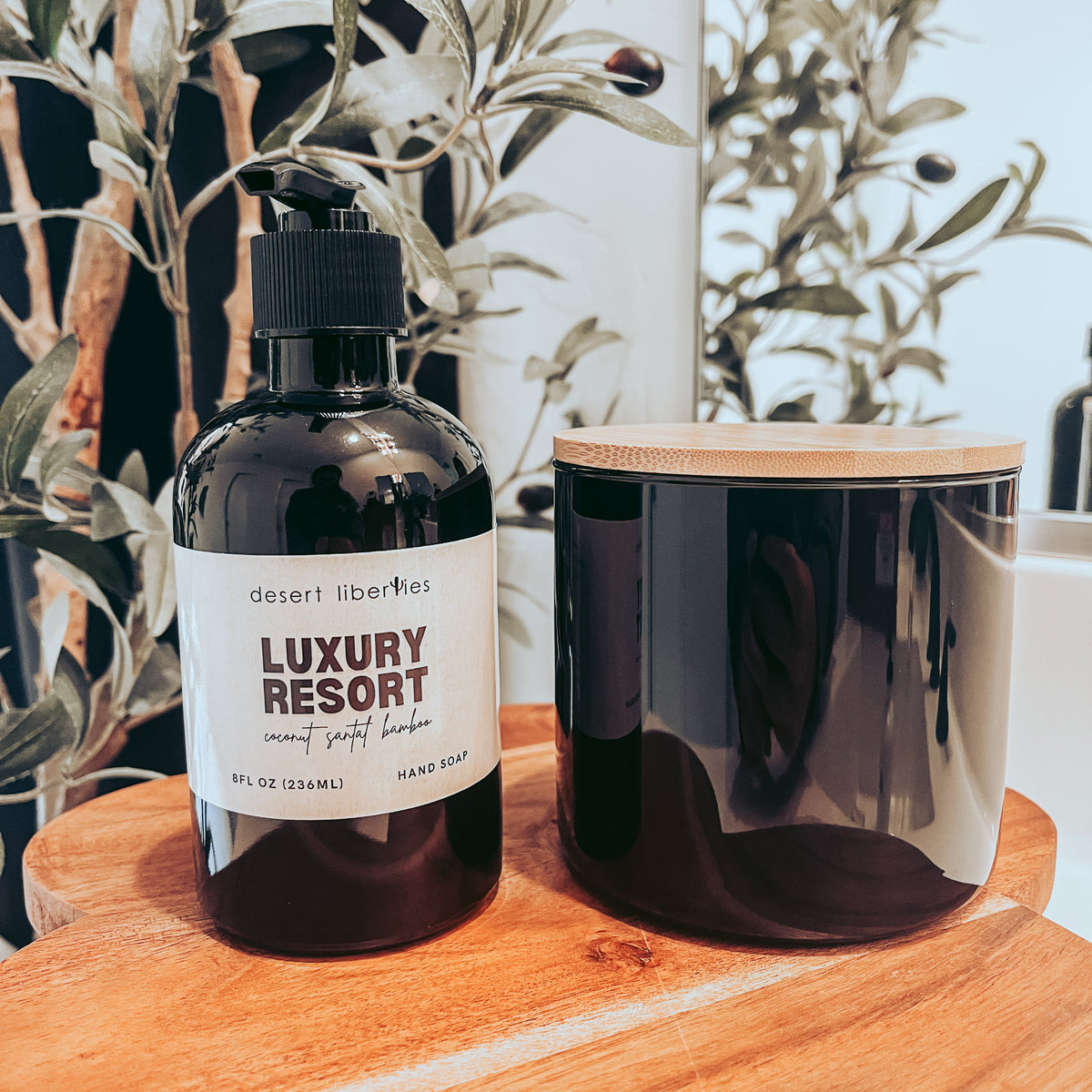 Soap bottle next to an unlabeled candle on a wood surface with olive leaves in the background.