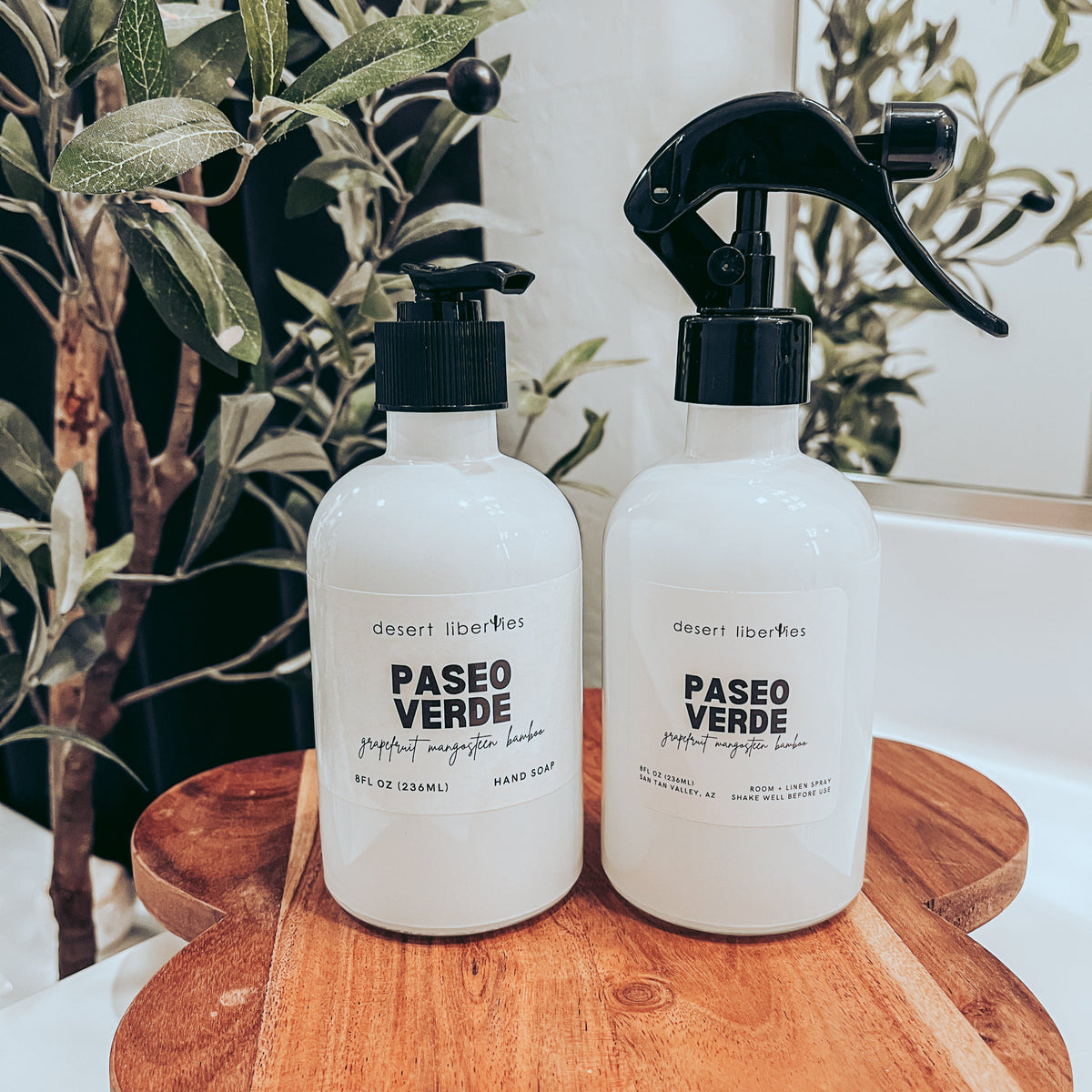 Soap bottle next to a matching spray bottle on a wood surface and olive tree in the background.