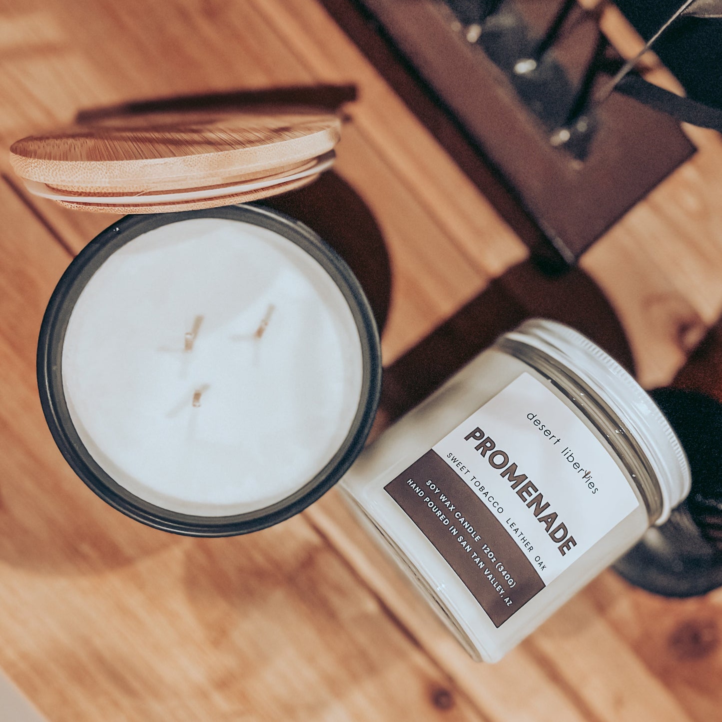 Top view of two candles, one with a bamboo lid and the other with a glass lid, both with white wax and labeled as PROMENADE. The candles are placed on a wooden surface.