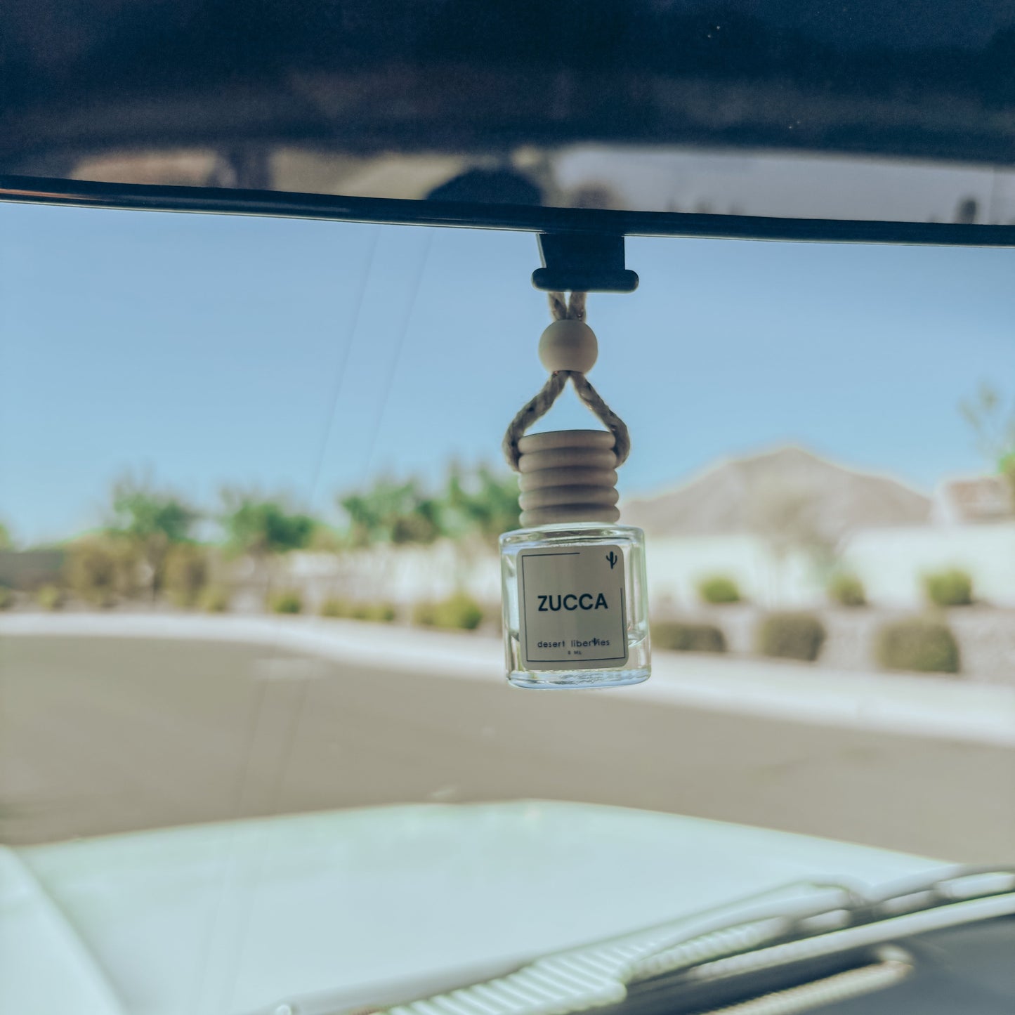 Zucca car air freshener hanging from a car's rearview mirror with a desert landscape in the background.