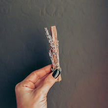 Load image into Gallery viewer, Hand holding a small bundle of dried lavender and palo santo against a textured gray wall.
