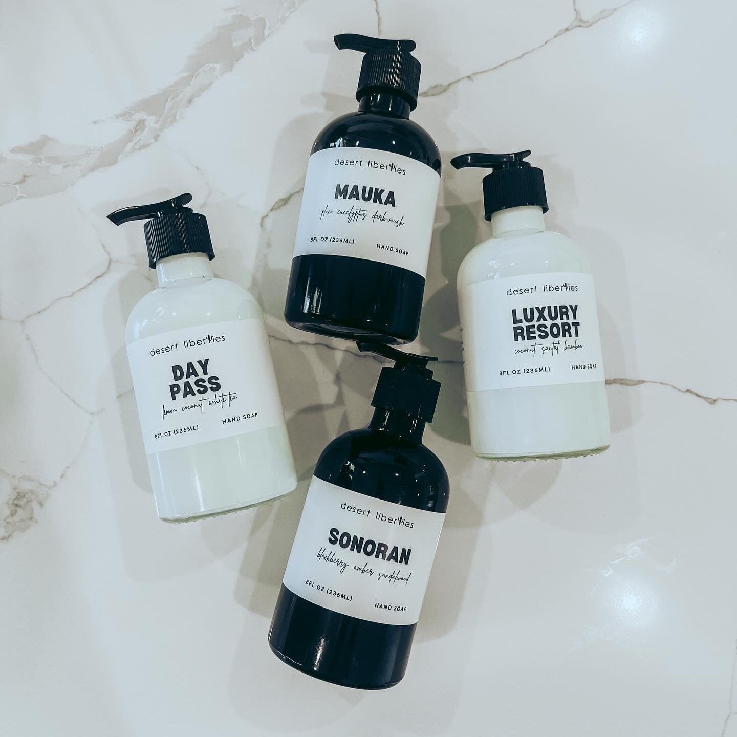 Four reusable glass bottles of organic hand soap with black pump dispensers, labeled with different scents such as 'DAY PASS', 'MAUKA', 'LUXURY RESORT', and 'SONORAN'. Two bottles are white and two are black, displayed on a marble surface.