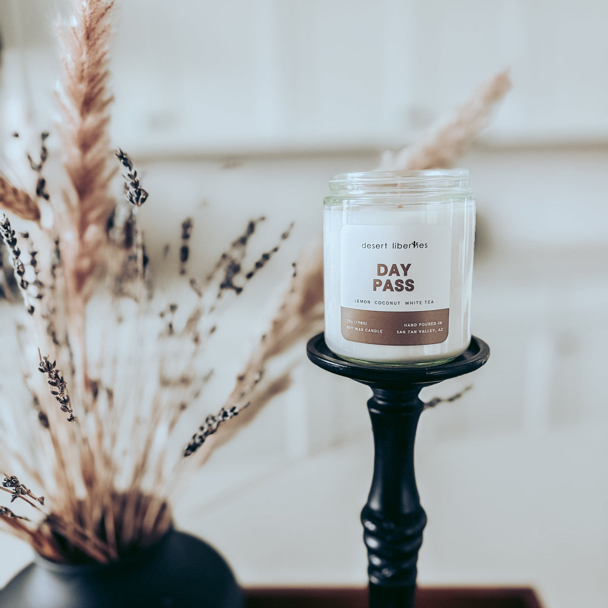 A white soy candle in a clear glass jar with a brown and white label displayed on a black candlestick holder. The scene is styled with a black vase of dried grasses in the foreground against a soft, blurred white paneled background.