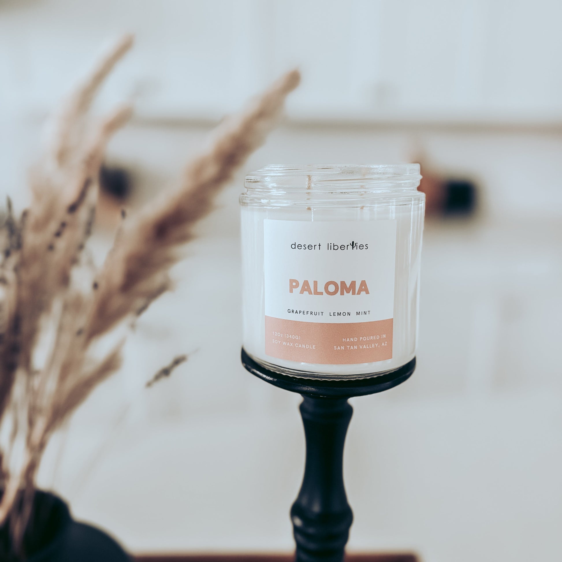 Candle labeled 'Paloma' on a stand with dried plants in the background