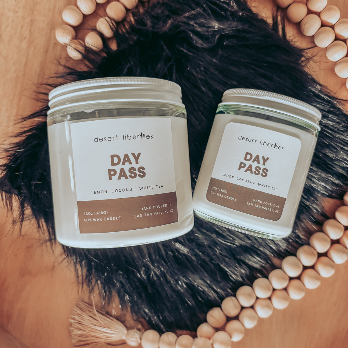 Two Desert Liberties 'Day Pass' scented candles in glass jars, styled on a wooden tray with black faux fur, wooden beads, and tassels.