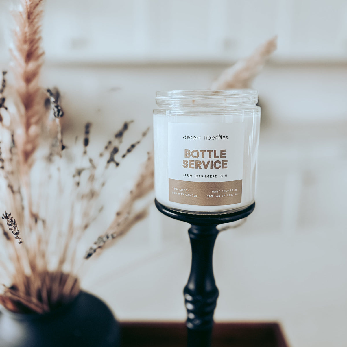 A clear glass jar candle labeled 'BOTTLE SERVICE' sits on a black candlestick holder on a wooden surface, with dried grasses and a dark vase blurred in the background.