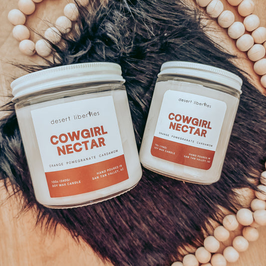 Two 'Cowgirl Nectar' candles on a fur surface with decorative beads and feathers.