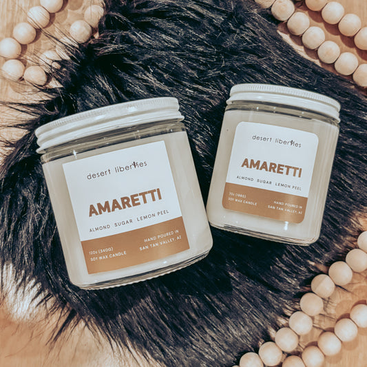 Two 'Amaretti' candles from Desert Liberties on a wooden surface with decorative beads.
