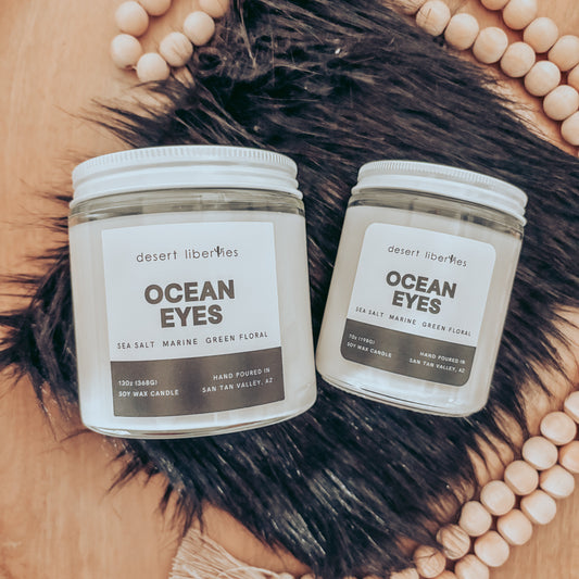 Two 'Ocean Eyes' candles on a wooden surface with decorative beads and fur.