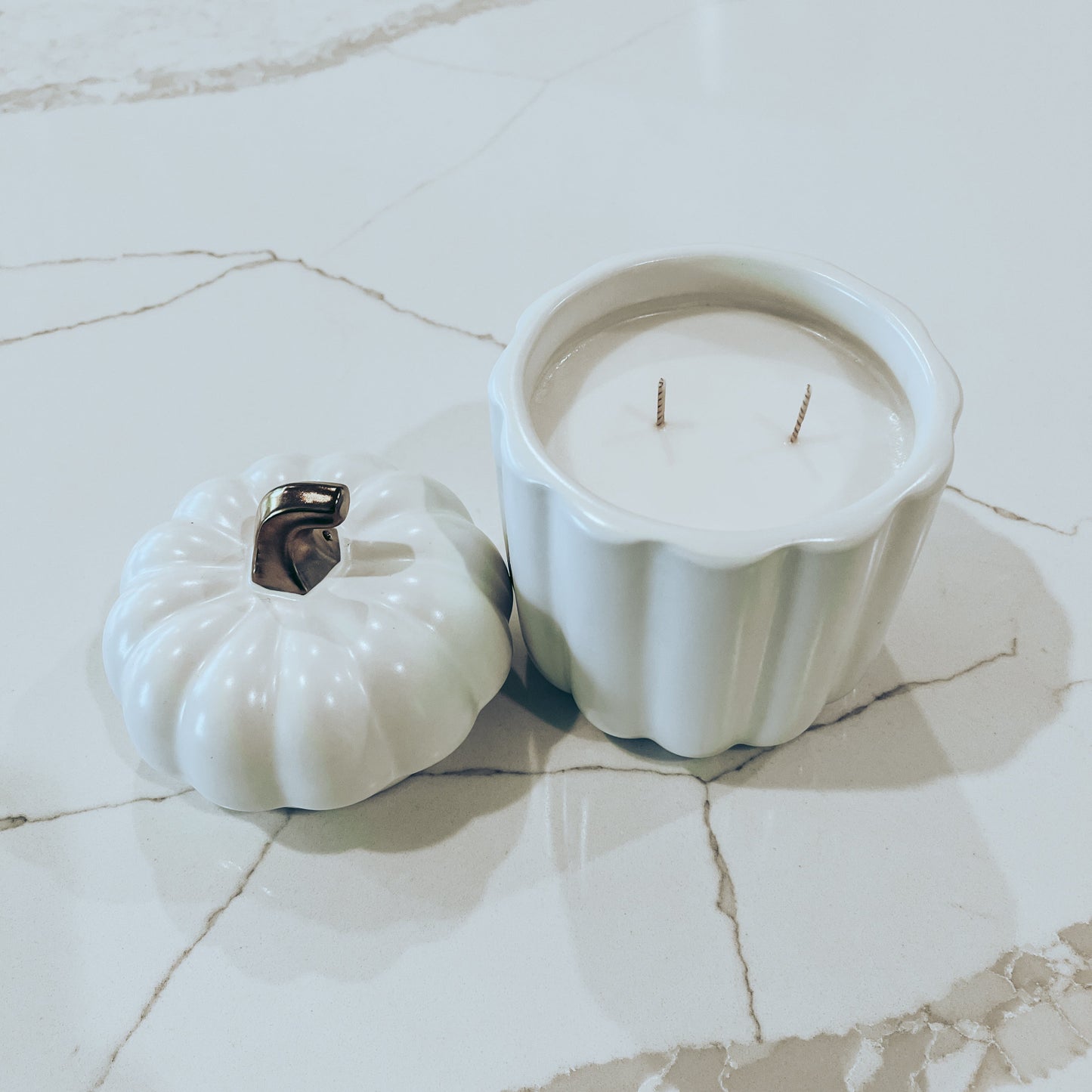 White and gold pumpkin canister candle, with top off, on a white marble surface. 