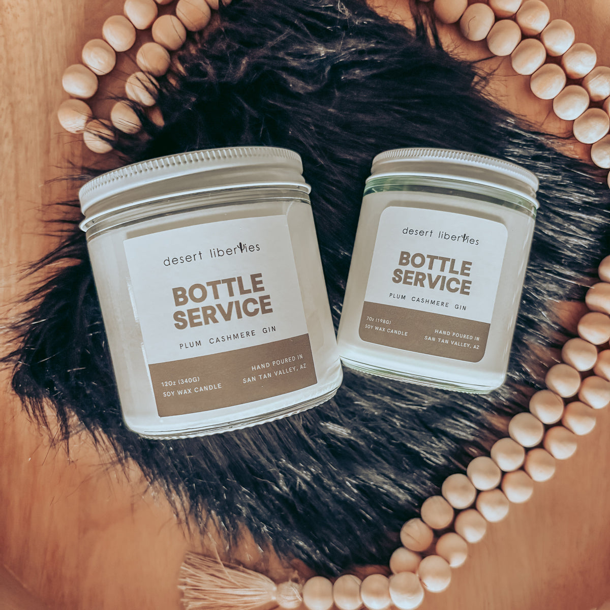 Two 'Bottle Service' scented candles by desert liberites are displayed in clear glass jars with white lids, arranged on a black faux fur rug inside a wooden bowl, surrounded by a wooden bead garland.
