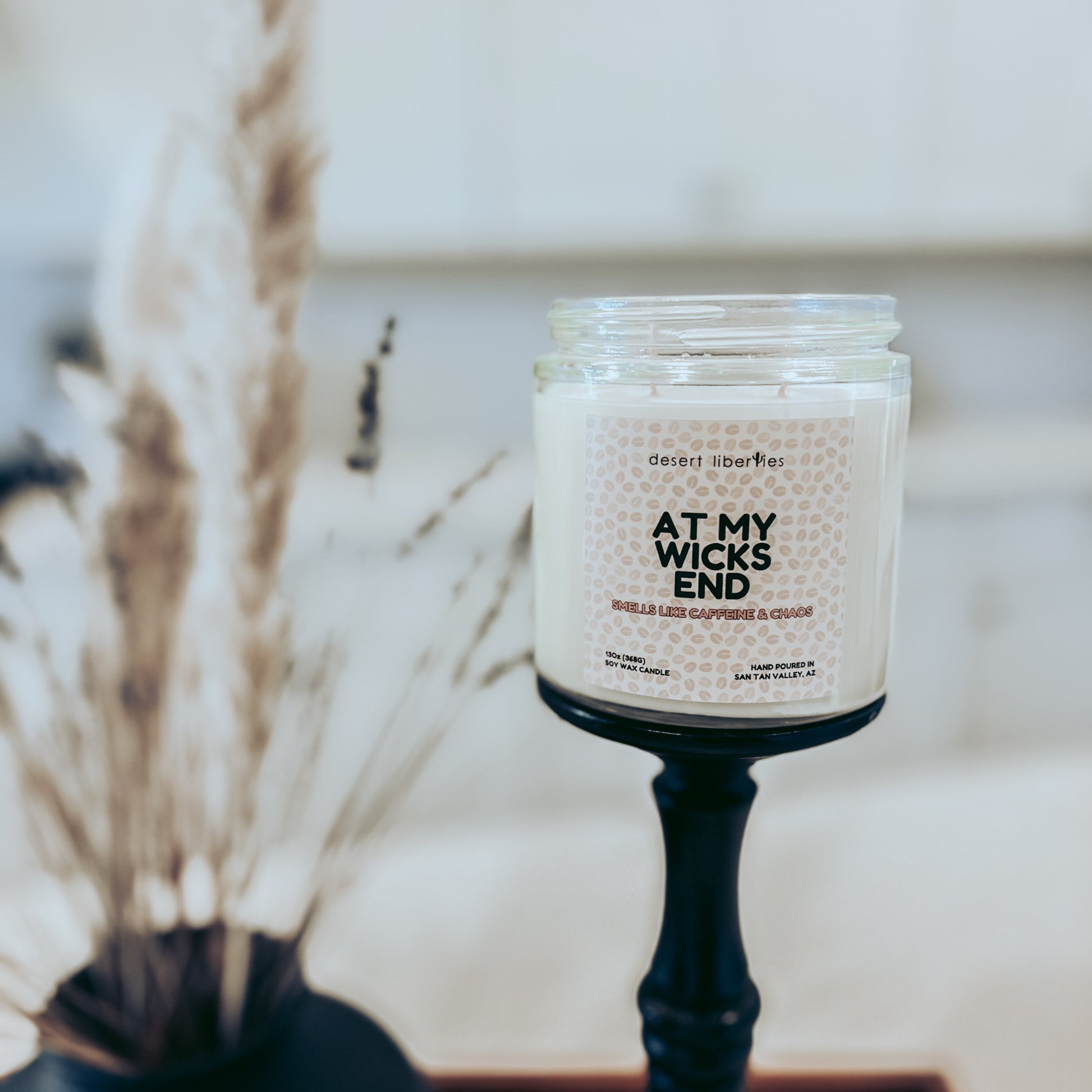 Candle labeled 'At My Wicks End' on a stand with dried plants in the background