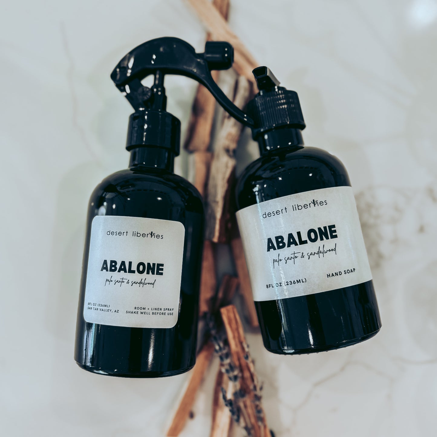 Two black bottles labeled 'Abalone' on a marble surface with palo santo sticks.