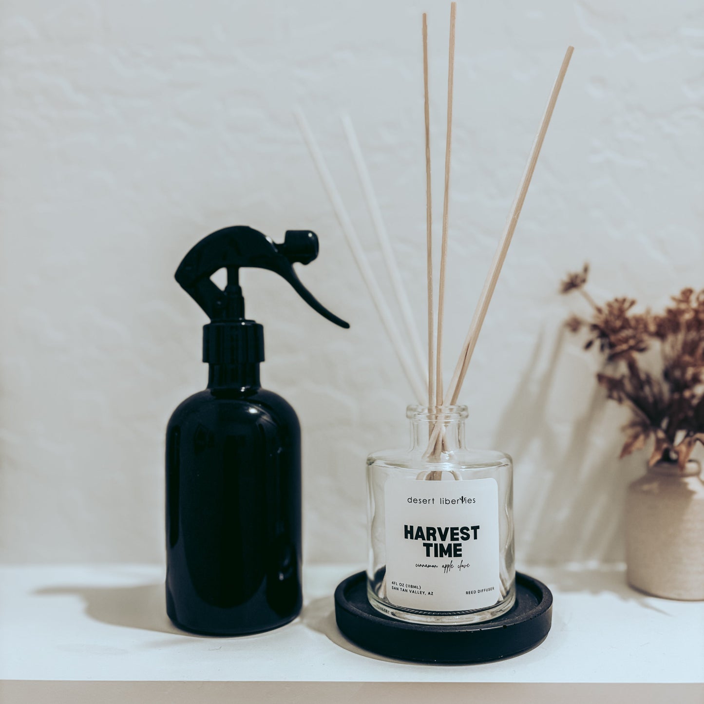 Black spray bottle and 'Harvest Time' reed diffuser with reeds on a white surface.