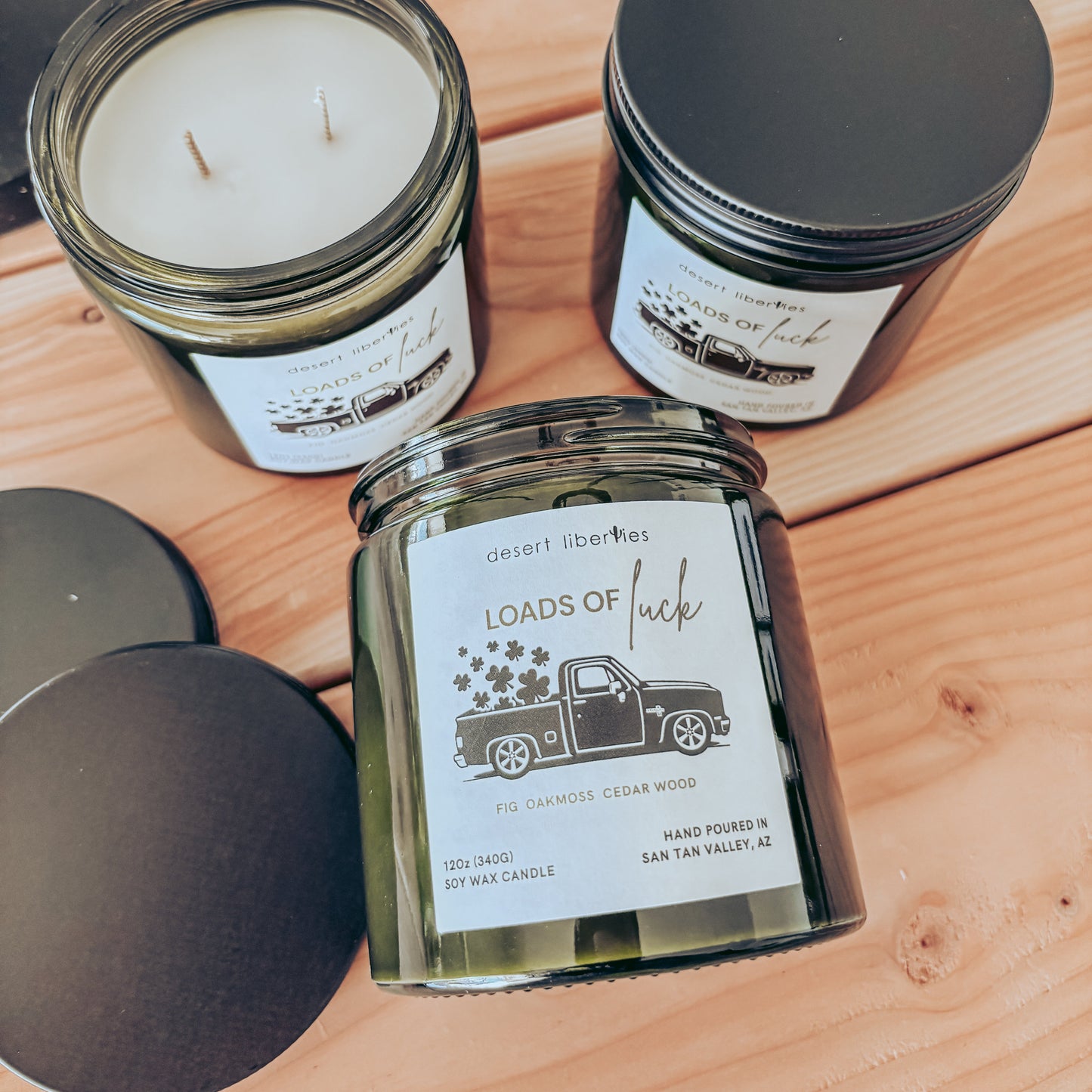 Three candles in glass jars with black lids on a wooden surface, featuring labels with truck graphics.