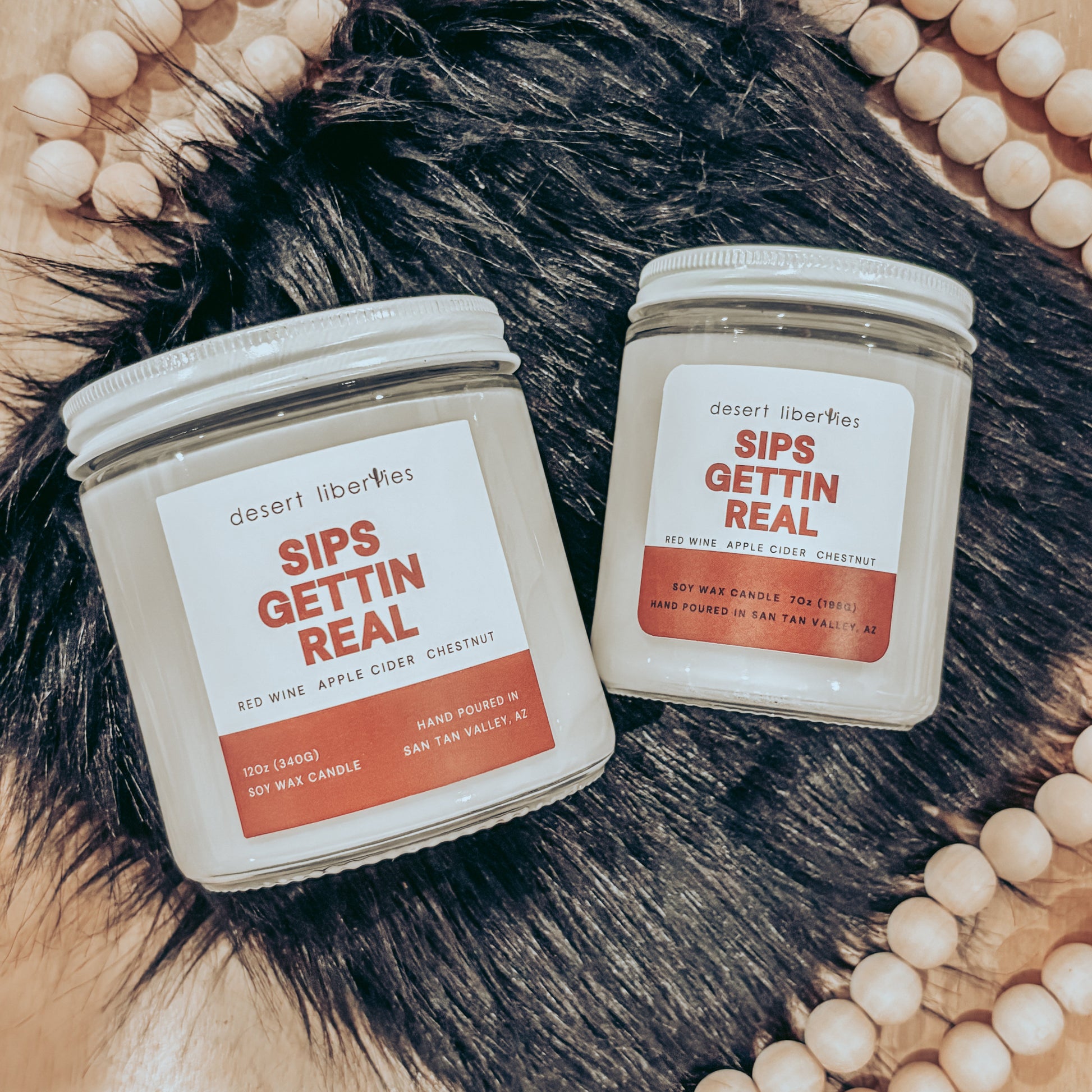 Two candles labeled 'Sips Gettin Real' on a fur surface with a beaded frame.
