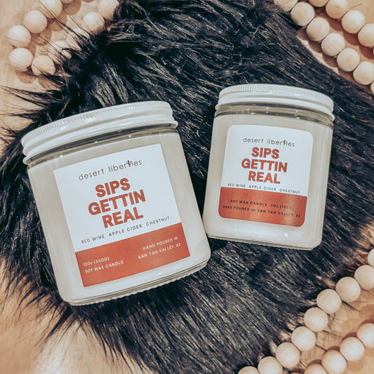 Two candles labeled 'Sips Gettin Real' on a fur surface with a beaded frame.