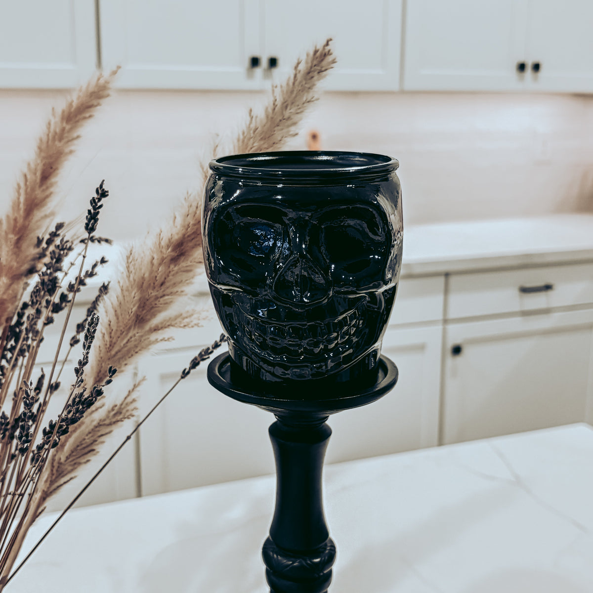 A black ceramic skull-shaped candle holder on a matching black candlestick stand, styled on a white table with dried pampas grass in a bright kitchen setting.