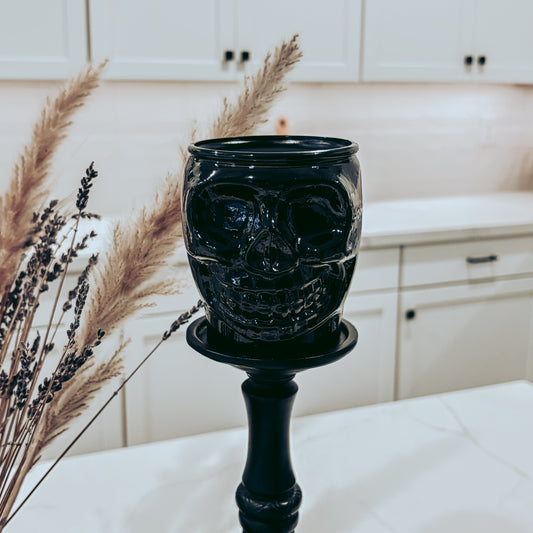 A black ceramic skull-shaped candle holder on a matching black candlestick stand, styled on a white table with dried pampas grass in a bright kitchen setting.