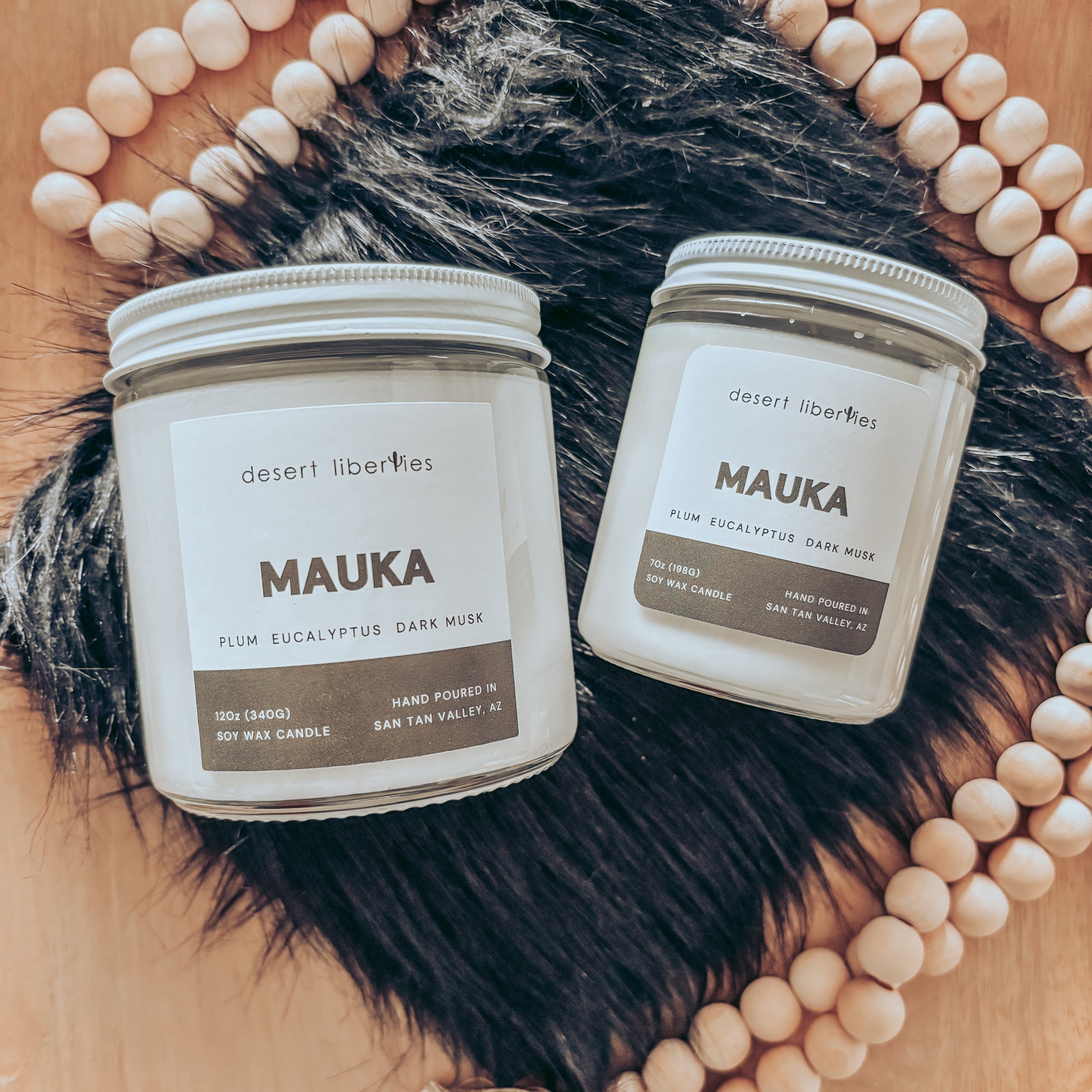 Two 'MAUKA' candles from Desert Liberties on a fur surface with decorative beads.