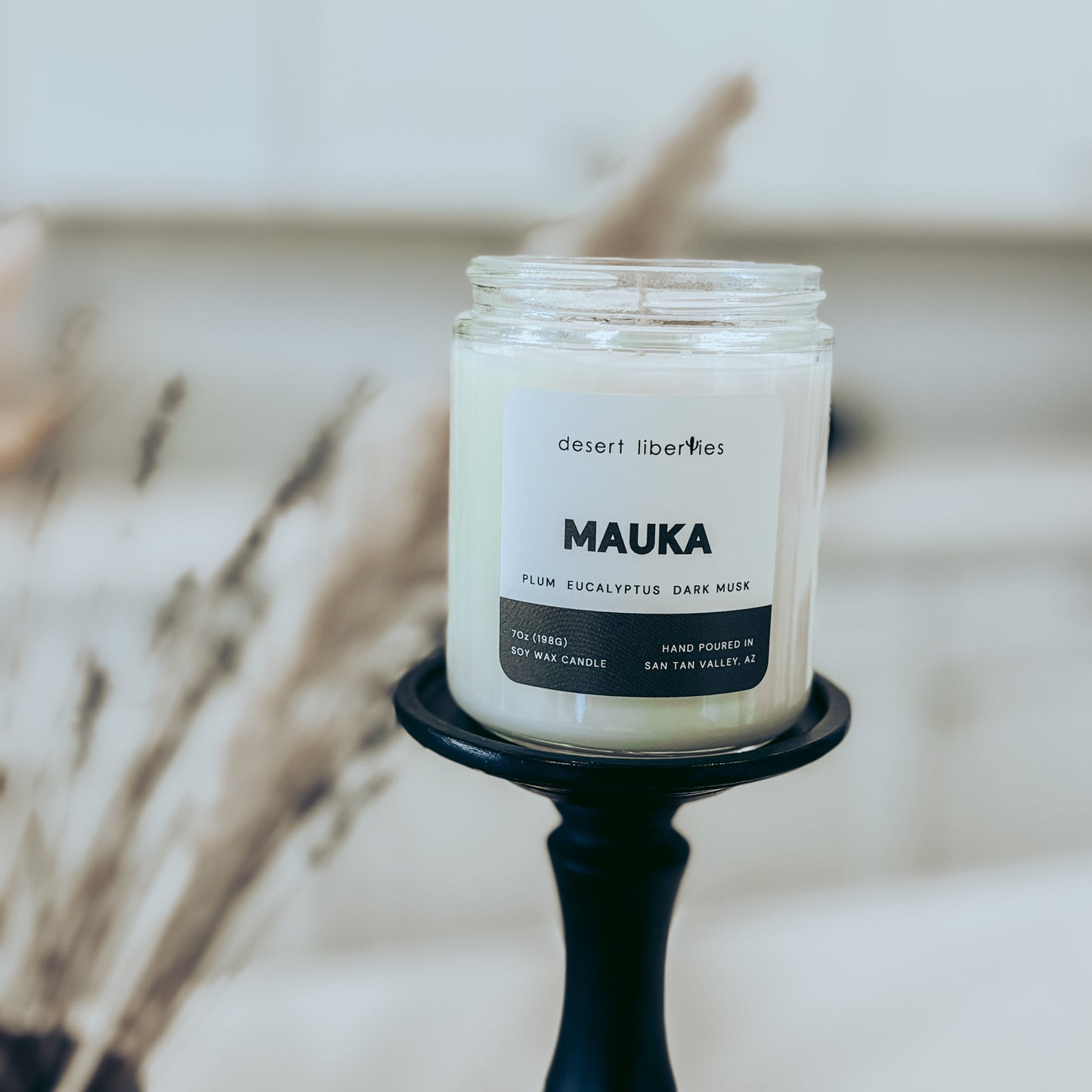 Candle in a glass jar labeled 'MAUKA' on a black holder with a blurred background