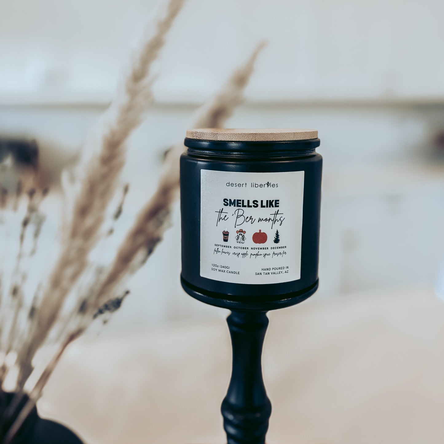 Black candle jar with wooden lid on a stand, featuring a label against a blurred natural background.