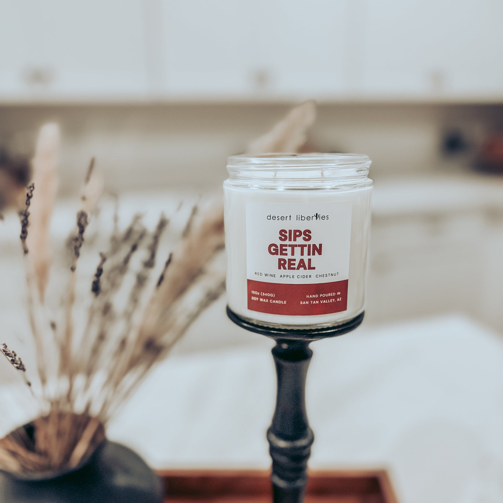 Candle with a label reading 'Sips Gettin Real' on a stand with decorative elements.