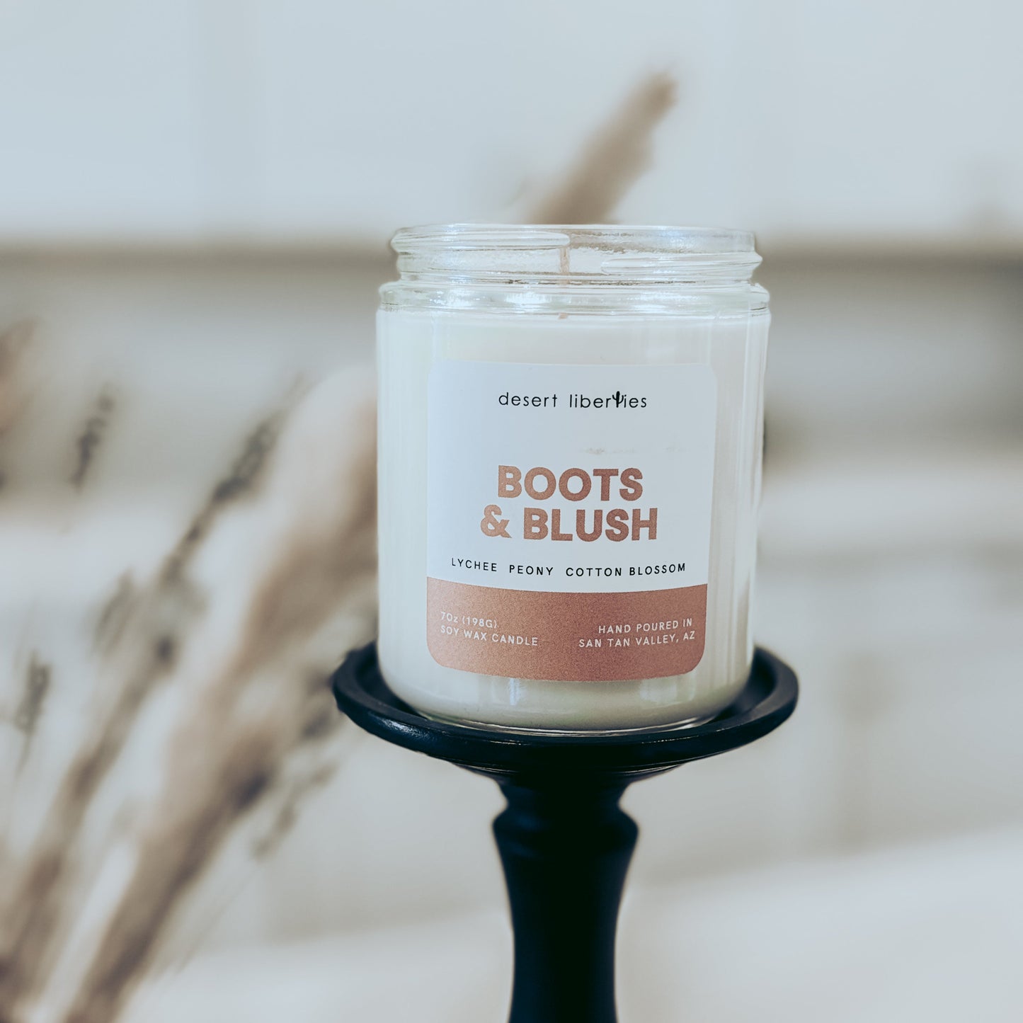 Candle in a jar labeled 'Boots & Blush' on a black candle holder with a blurred background.