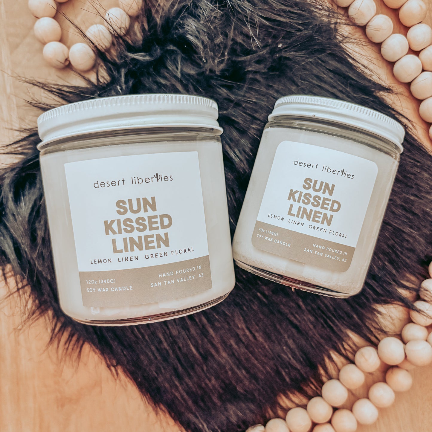 Two 'Sun Kissed Linen' candles by Desert Liberties on a textured surface with beads and feathers.