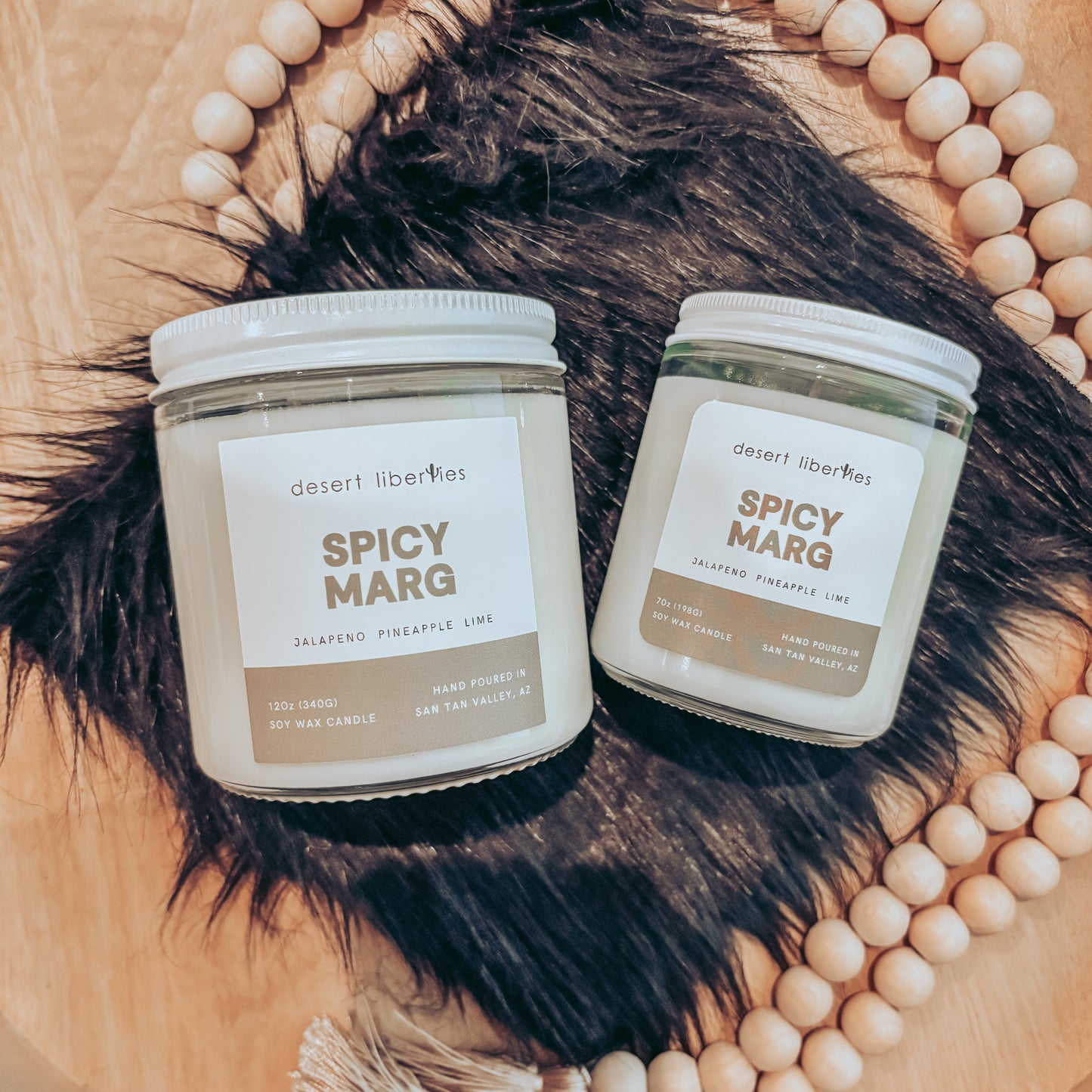 Two candles labeled 'Spicy Marg' on a wooden surface with decorative beads and fur.