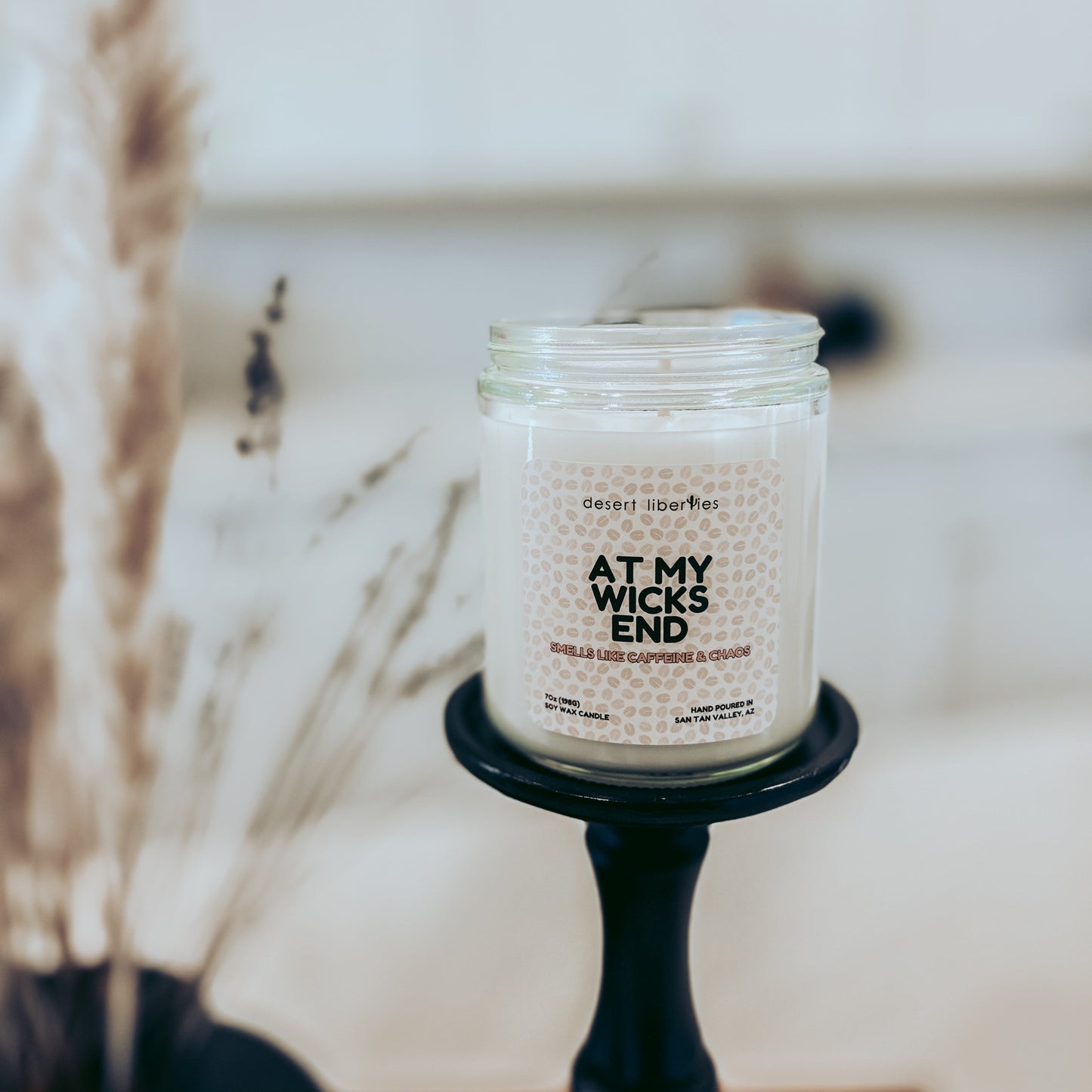 Candle in a glass jar labeled 'At My Wicks End' on a black candlestick with a blurred background.