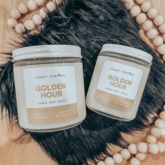 Two candles with white labels, one small and one medium, in clear glass jars. The labels say 'GOLDEN HOUR' and list fragrance notes.