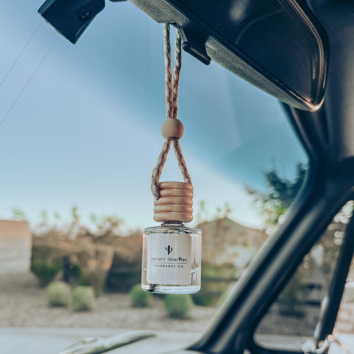 Glass diffuser bottle hanging from a rear view mirror in a car with desert background blurred. 