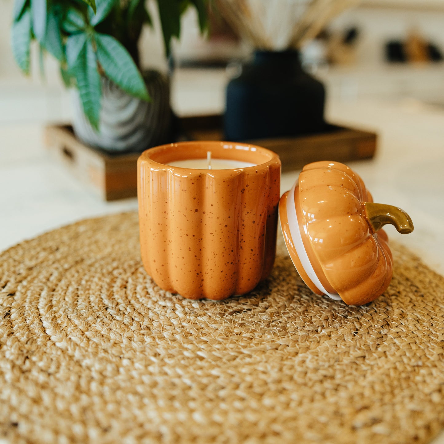 Pumpkin-shaped candle on a woven mat with a plant and kitchen items in the background