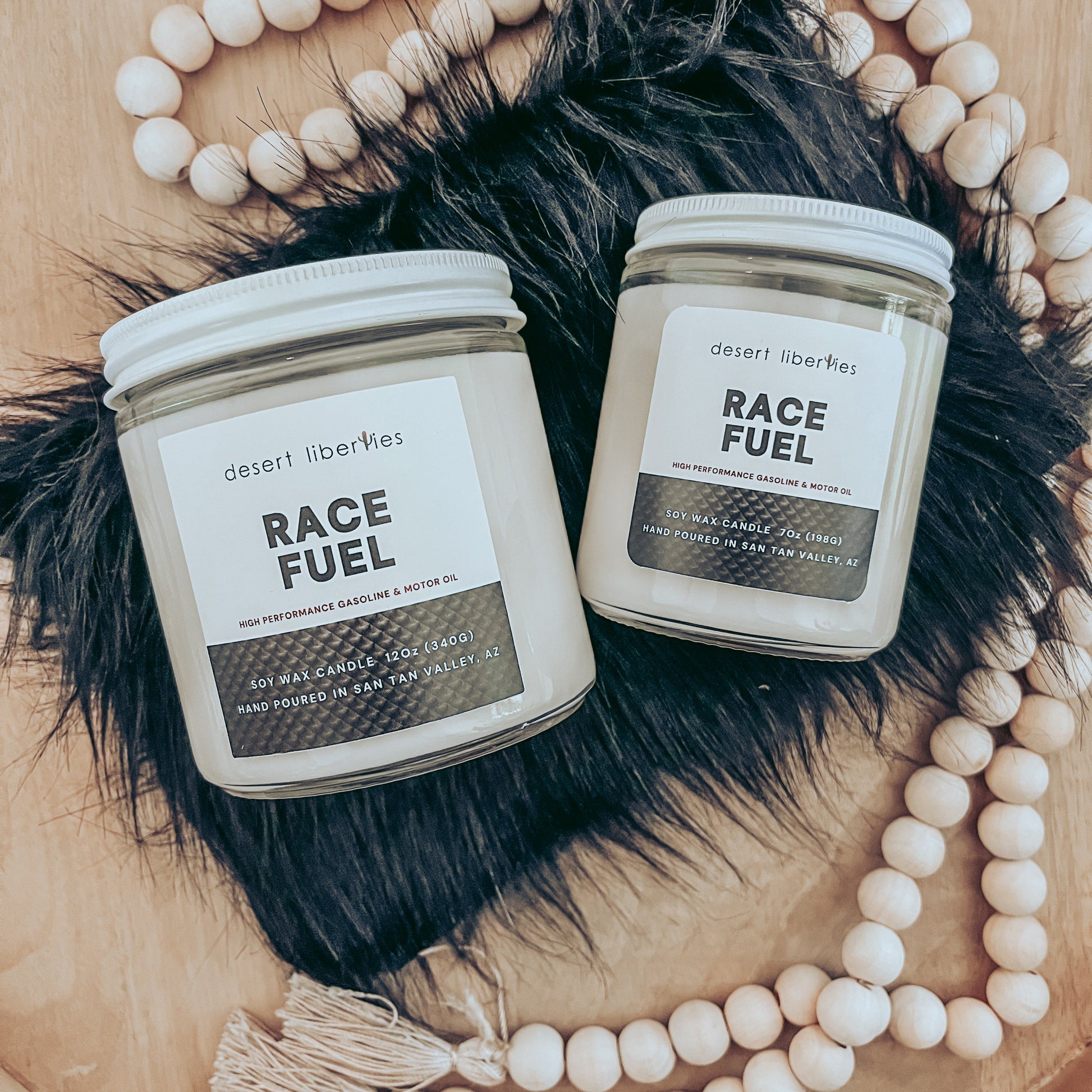 Two candles labeled 'RACE FUEL' by Desert Liberties, with carbon fiber-like label, placed on a wooden surface with a black fur accent and beads, with a shirt in the background.