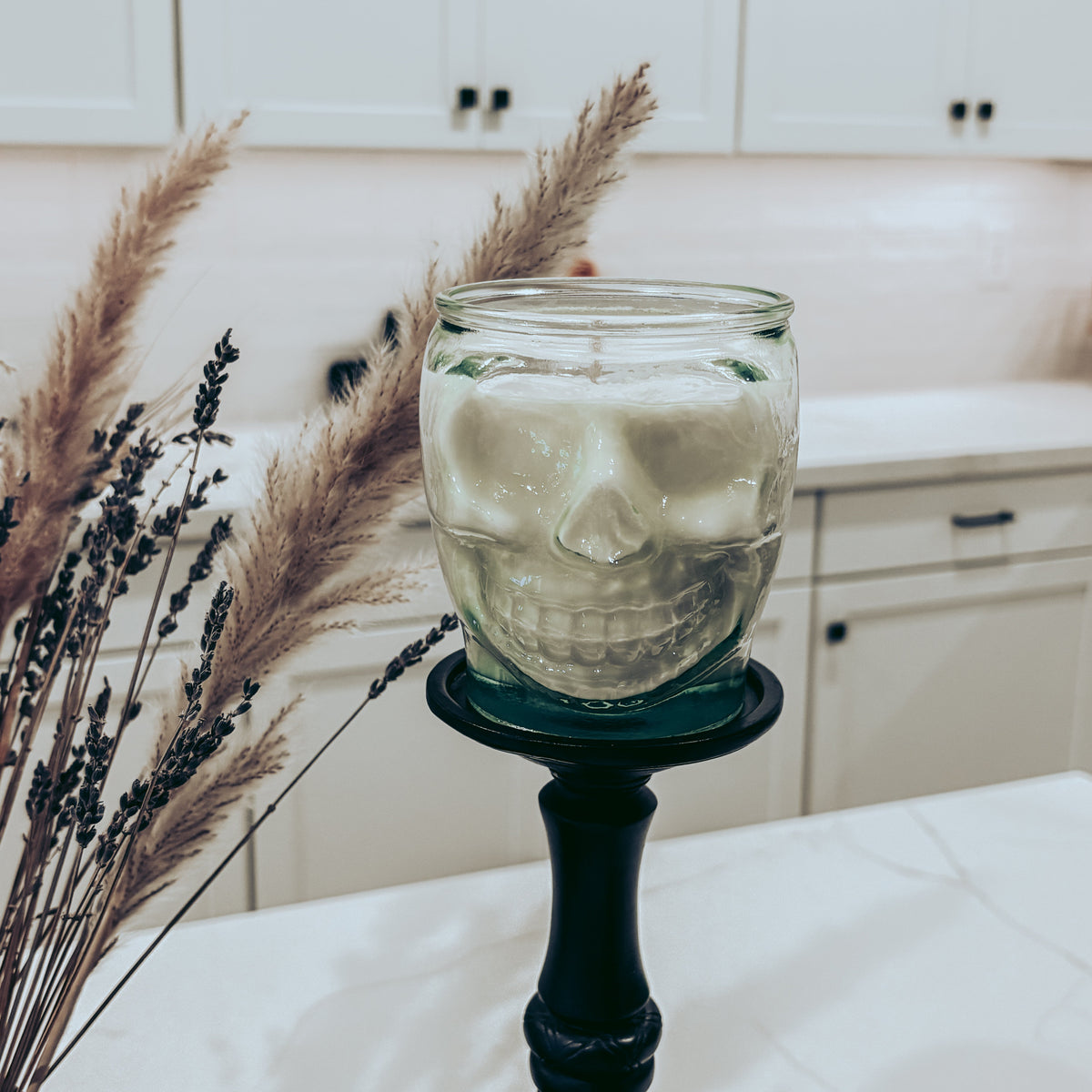 A skull-shaped glass jar candle filled with white wax, displayed on a black candlestick holder on a kitchen countertop with dried pampas grass in the foreground.