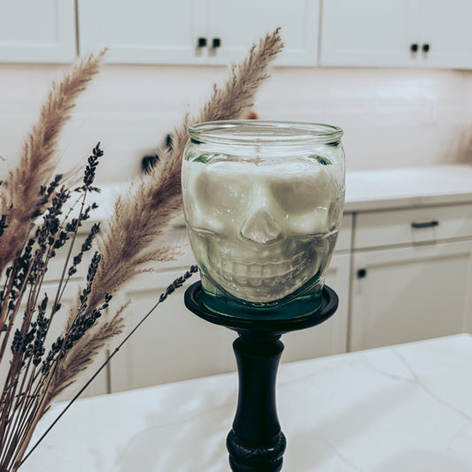 A skull-shaped glass jar candle filled with white wax, displayed on a black candlestick holder on a kitchen countertop with dried pampas grass in the foreground.