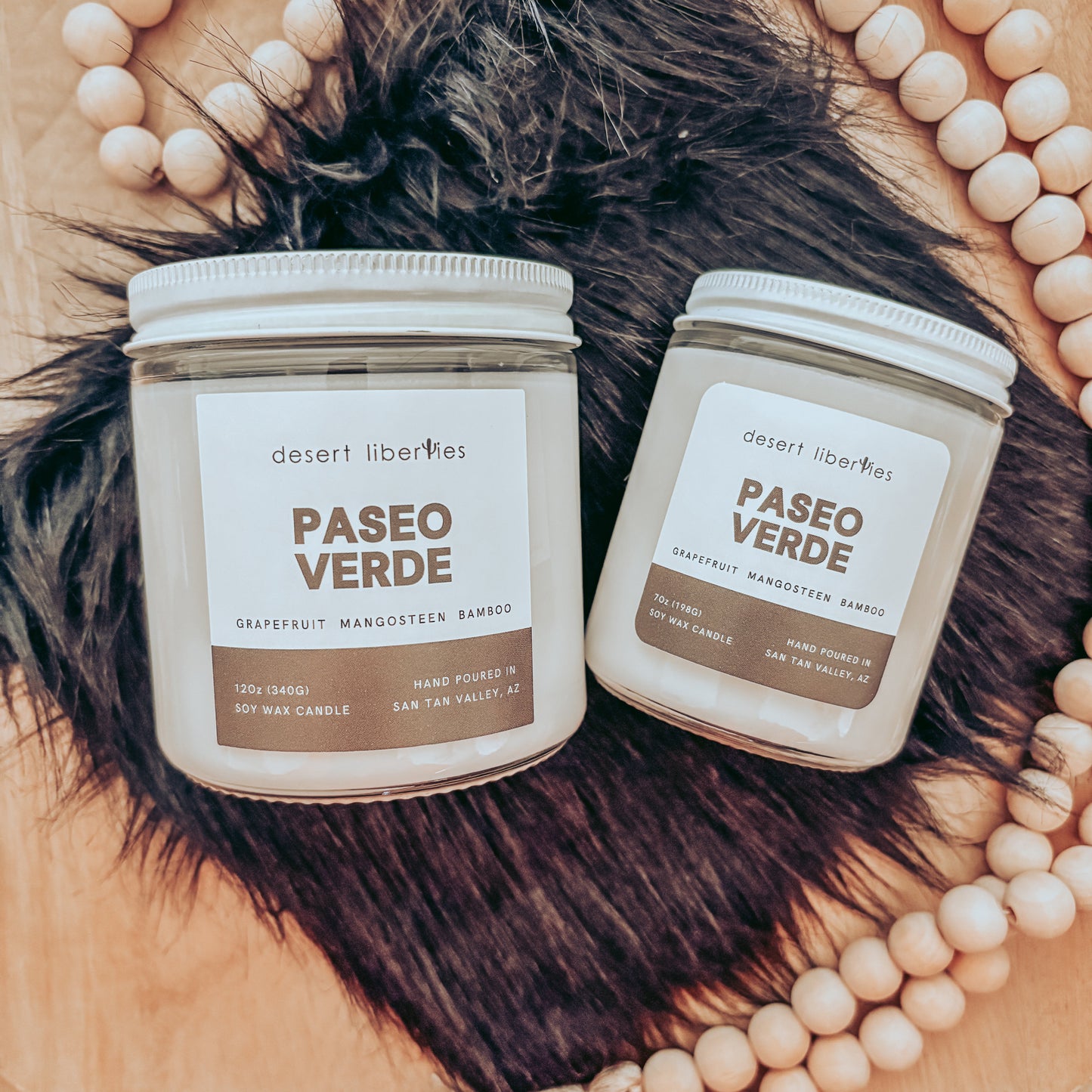 Two candles labeled 'Paseo Verde' on a textured surface with decorative beads and a hat in the background.