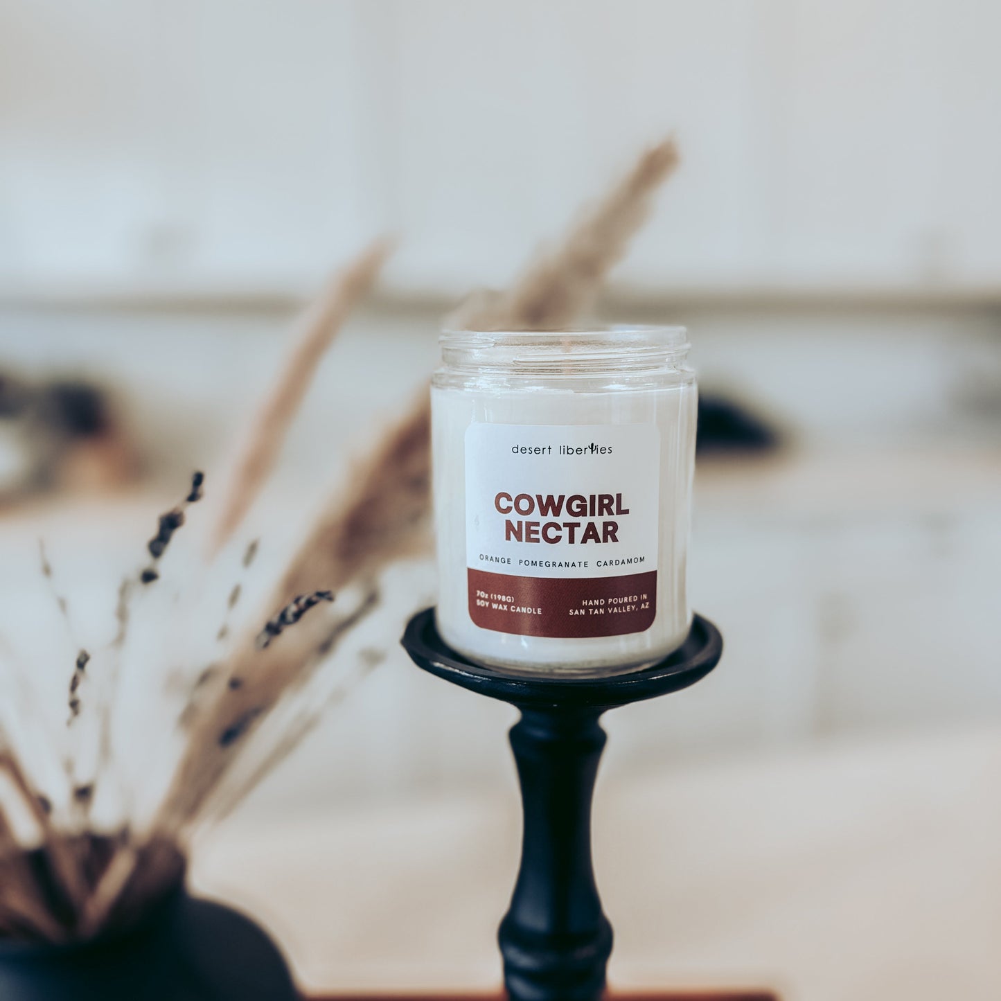 Candle labeled 'Cowgirl Nectar' on a stand with dried plants in the background