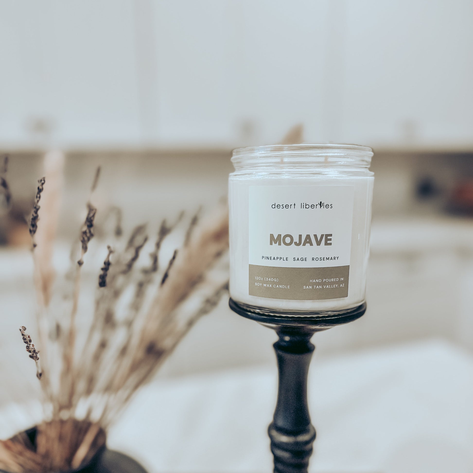 Candle with 'Mojave' label on a stand, surrounded by dried plants.